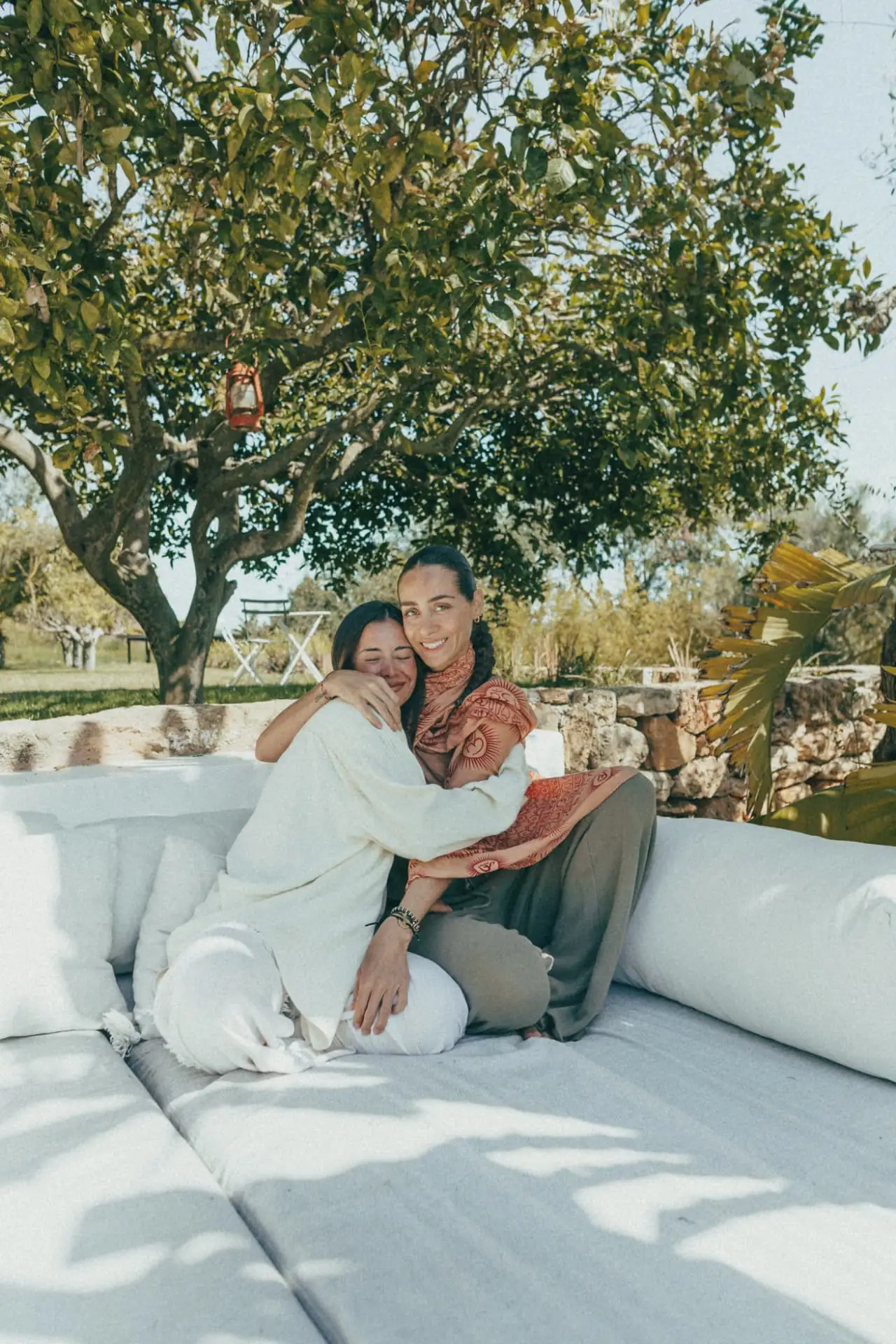 Two guests sitting close together on a garden daybed under a fruit tree